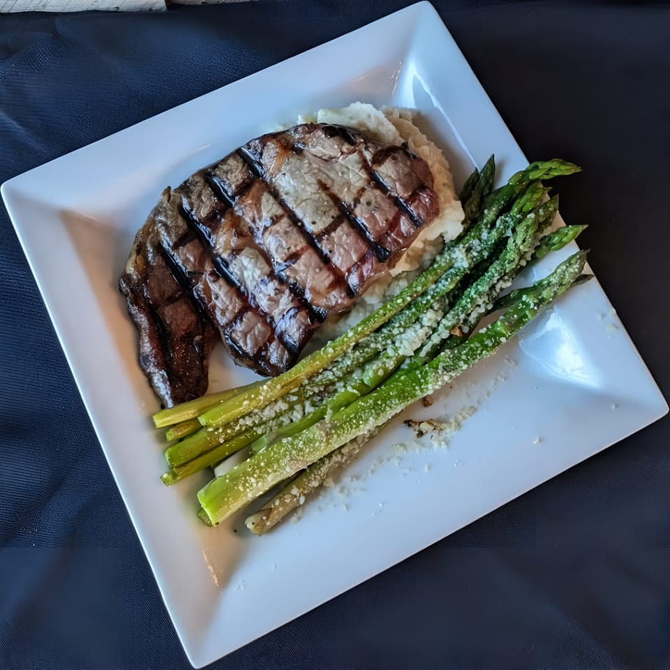 Grilled steak with mashed potatoes and parmesan-dusted asparagus on a white plate at Bucket List Tavern.