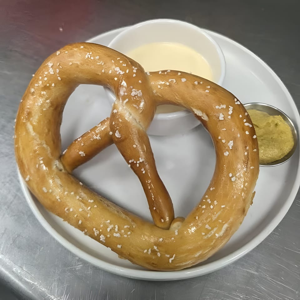 A large, salted pretzel with cheese dip and mustard on a white plate at Bucket List Tavern.