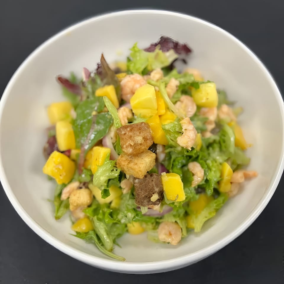A vibrant salad with green lettuce, yellow mango chunks, shrimp, and croutons in a white bowl.