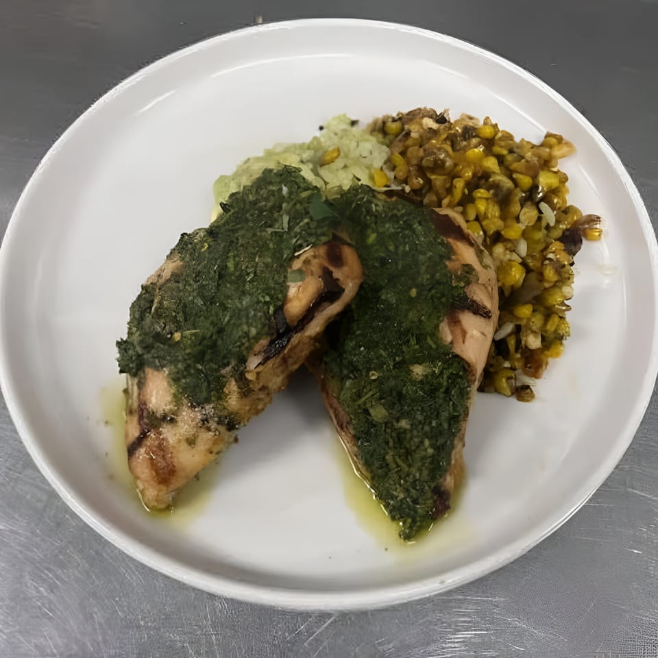 Grilled chicken breasts topped with green pesto, served with corn and rice on a white plate.