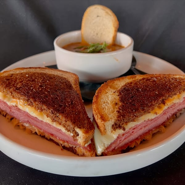 A grilled Reuben sandwich, cut in half, served with a side of tomato soup and a bread slice.